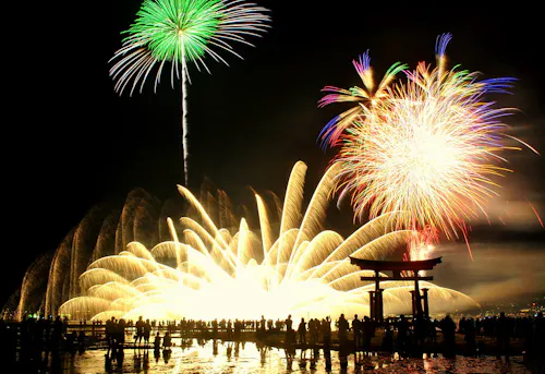 A crowd watches vibrant fireworks illuminating the night sky above a traditional Japanese torii gate, with bright green, gold, and multicolored bursts reflecting on water.