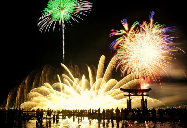 A crowd watches vibrant fireworks illuminating the night sky above a traditional Japanese torii gate, with bright green, gold, and multicolored bursts reflecting on water.