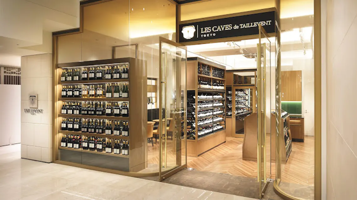 The image shows the entrance to "Les Caves de Taillevent" in Tokyo, a well-lit wine shop with numerous bottles displayed in wooden shelves lining the walls. Glass doors are open, inviting customers inside. The interior features sleek wood and modern design elements.