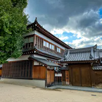 Daigan-ji Temple Traditional Japanese building with wooden exterior and grey tiled roof set against a cloudy sky. A large tree stands to the left, and a sandy path runs in front of the structure.