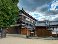 Traditional Japanese building with wooden exterior and grey tiled roof set against a cloudy sky. A large tree stands to the left, and a sandy path runs in front of the structure.