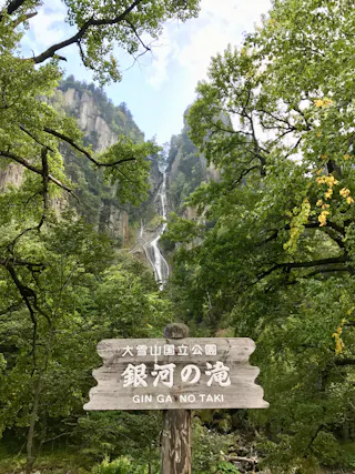 A wooden sign reading "GIN GA NO TAKI" stands among lush green trees, with a tall, narrow waterfall cascading down rocky cliffs in the background under a partly cloudy sky.