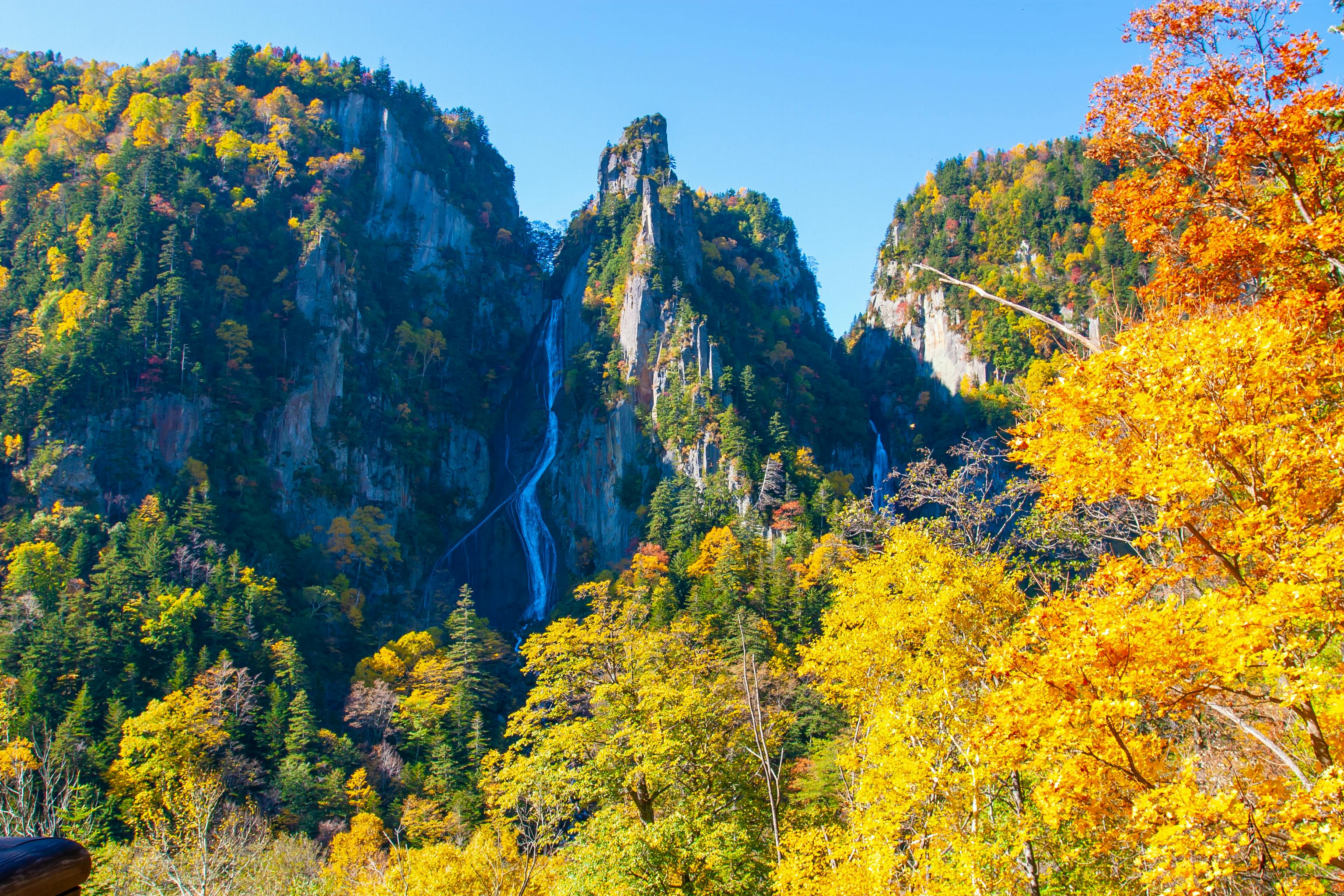 A tall waterfall flows down a rocky, forested mountain surrounded by vibrant autumn foliage in shades of yellow, orange, and green under a clear blue sky.