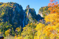 A tall waterfall flows down a rocky, forested mountain surrounded by vibrant autumn foliage in shades of yellow, orange, and green under a clear blue sky.
