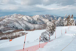 Snow-covered mountain landscape with pine trees and rolling hills in the background. Orange safety fencing and poles mark the edge of a skiing or snowboarding path on the snowy slope under a cloudy sky.