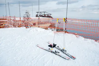 Two pairs of skis, ski poles, and gloves rest on the snow near an orange safety fence at a snowy mountain top, with trees and ski lift equipment visible in the background.