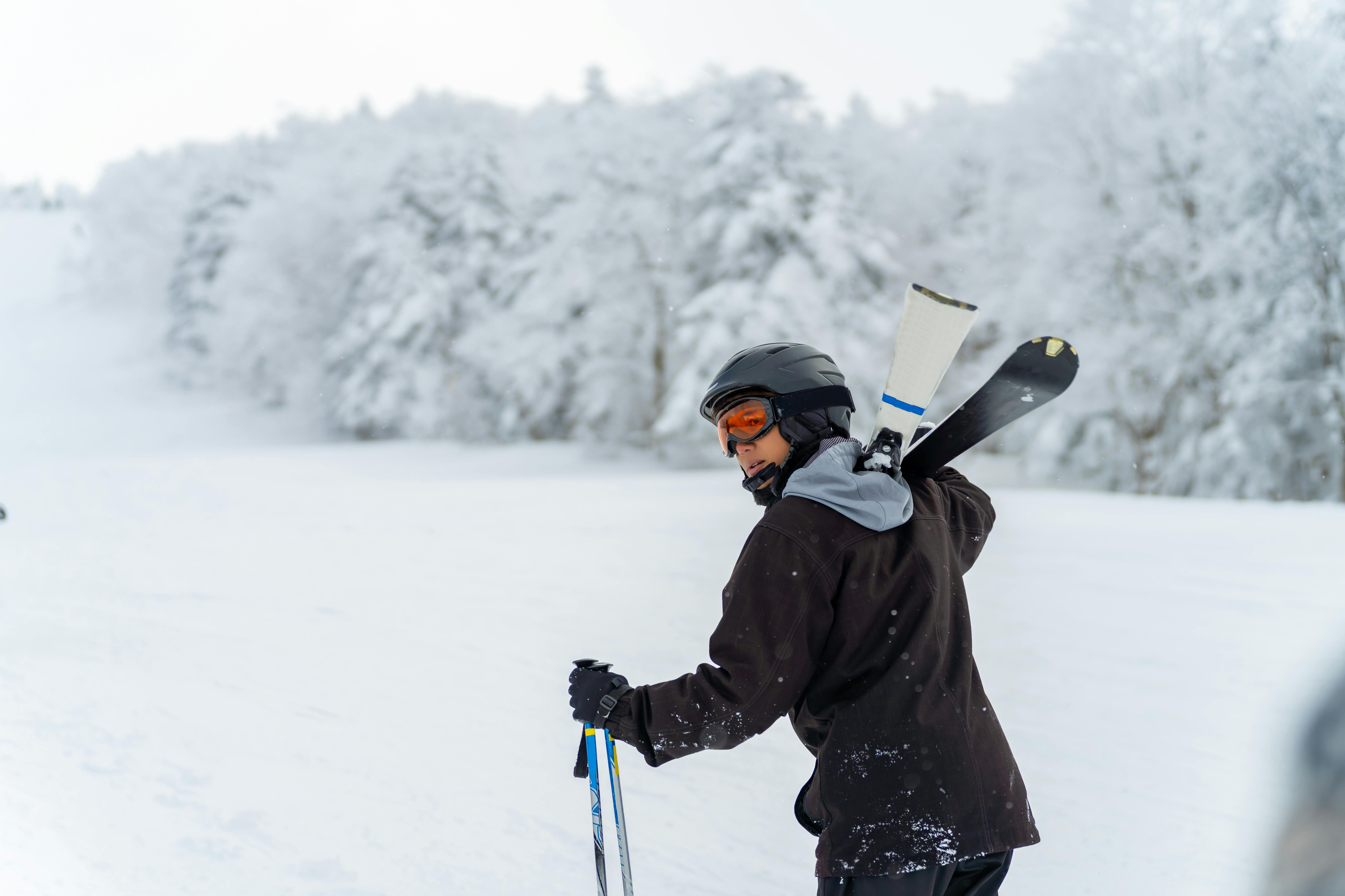 A skier wearing a helmet and goggles stands on a snowy slope, carrying skis over one shoulder and holding poles, with snow-covered trees in the background.
