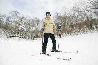 A person wearing a beige jacket, black pants, gloves, and sunglasses stands on skis holding ski poles on a snowy slope, with bare trees and a cloudy sky in the background.