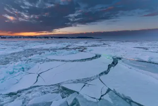 A frozen sea with large cracks in the ice stretches toward a distant shore under a dramatic sky at sunset, with hints of orange light on the horizon and dark clouds above.
