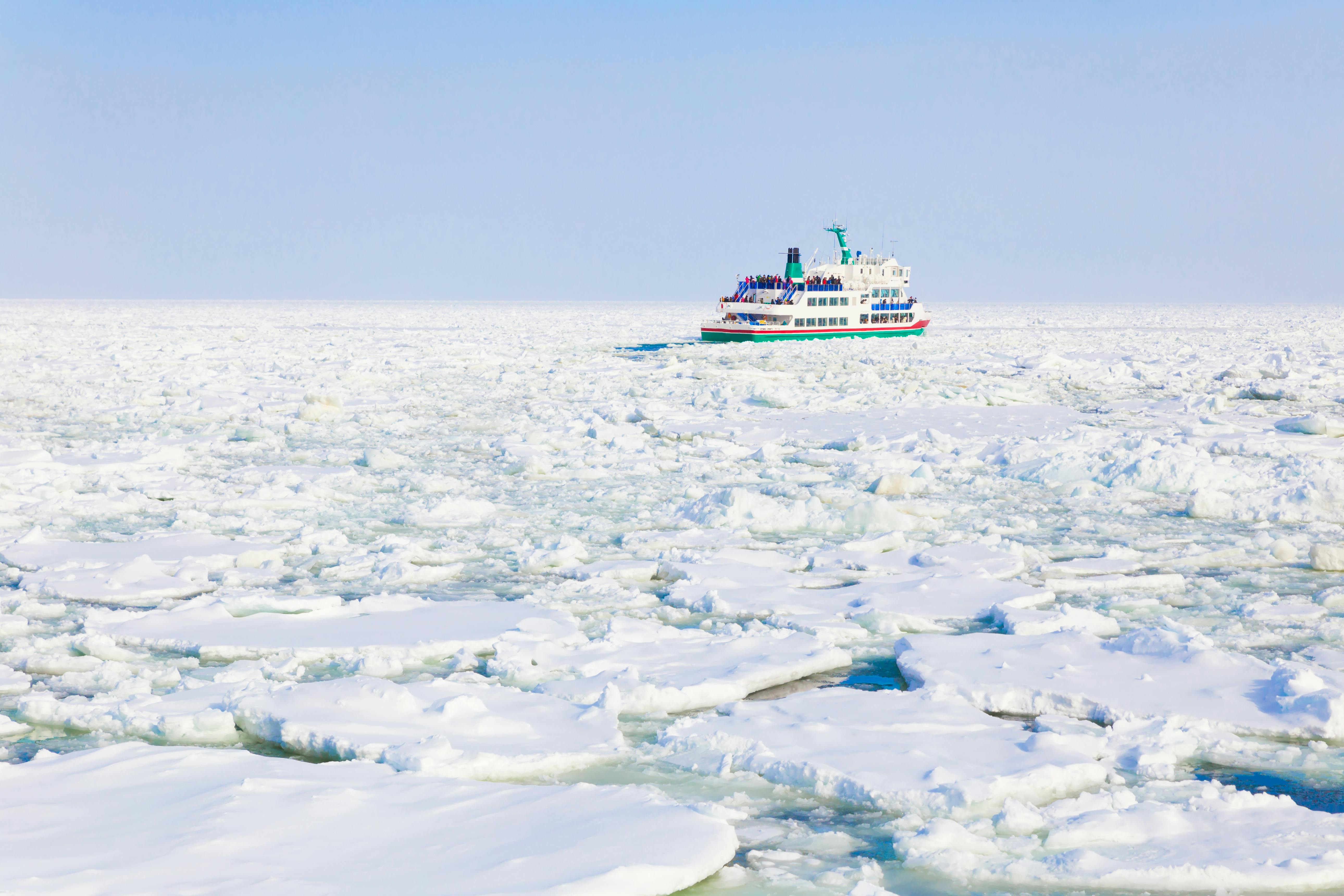 A white passenger ship travels through a vast, icy sea filled with floating sheets of ice under a clear blue sky. The boat is surrounded by snow and ice, with no land in sight.