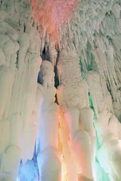 Large icicles and thick ice formations hang from a cave ceiling, illuminated with soft pastel lights in shades of pink, orange, blue, and green, creating a magical, surreal atmosphere.