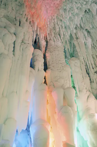 Large icicles and thick ice formations hang from a cave ceiling, illuminated with soft pastel lights in shades of pink, orange, blue, and green, creating a magical, surreal atmosphere.