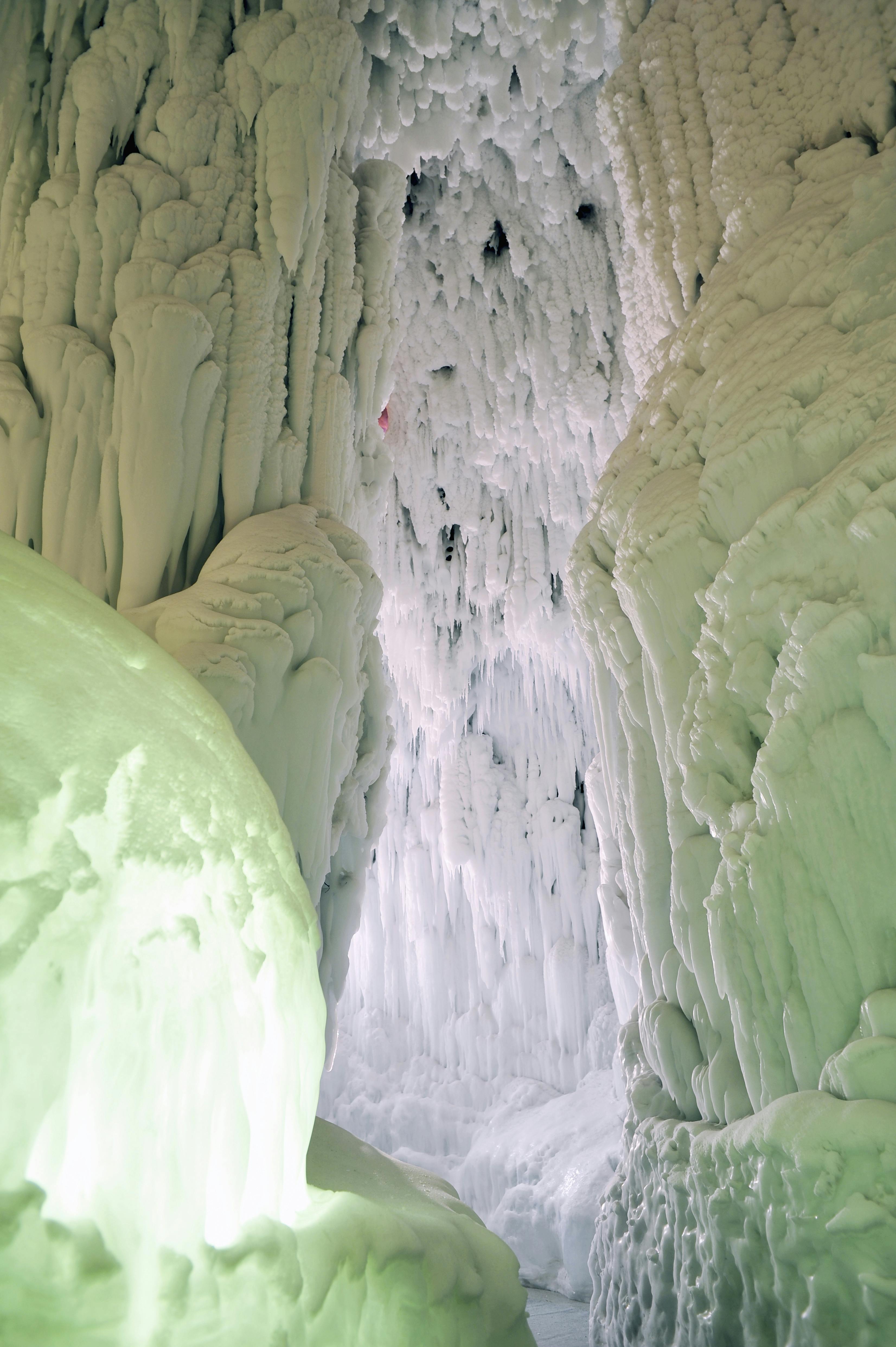 Narrow pathway between tall, thick walls of icicles and frozen formations, with white and pale green hues, creating an icy cave-like scene.