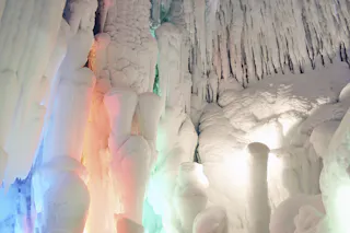 Icicles and frozen formations inside an ice cave are illuminated by colorful lights in shades of orange, green, and white, creating a glowing, magical winter scene.