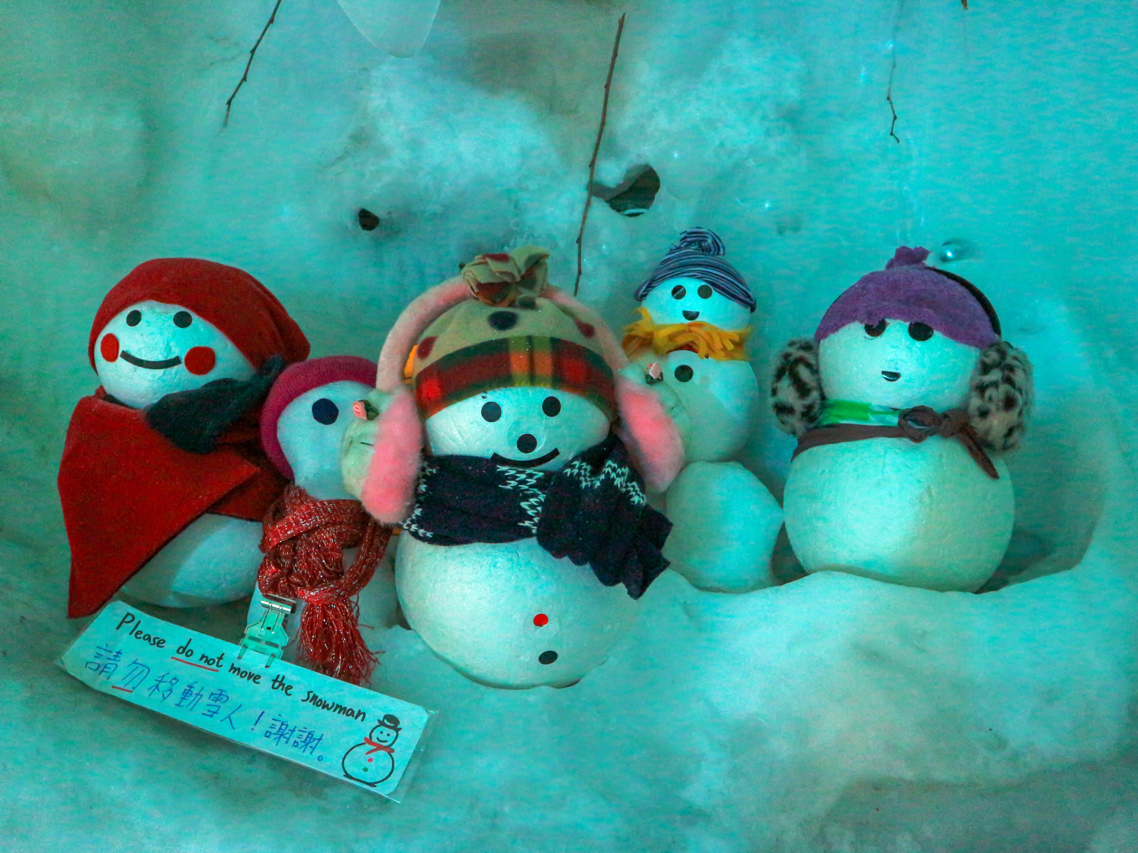 A group of six snowmen wearing colorful hats, scarves, and earmuffs sits together on snow. A sign in English and Japanese on the left reads, "Please do not move the snowman." The scene has a blue-tinted light.