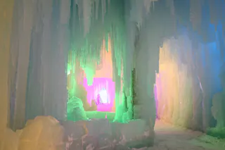 Large icicles and ice formations hang from the ceiling and walls of an ice cave, illuminated by colorful lights in shades of green, pink, purple, and yellow, creating a magical, glowing effect.