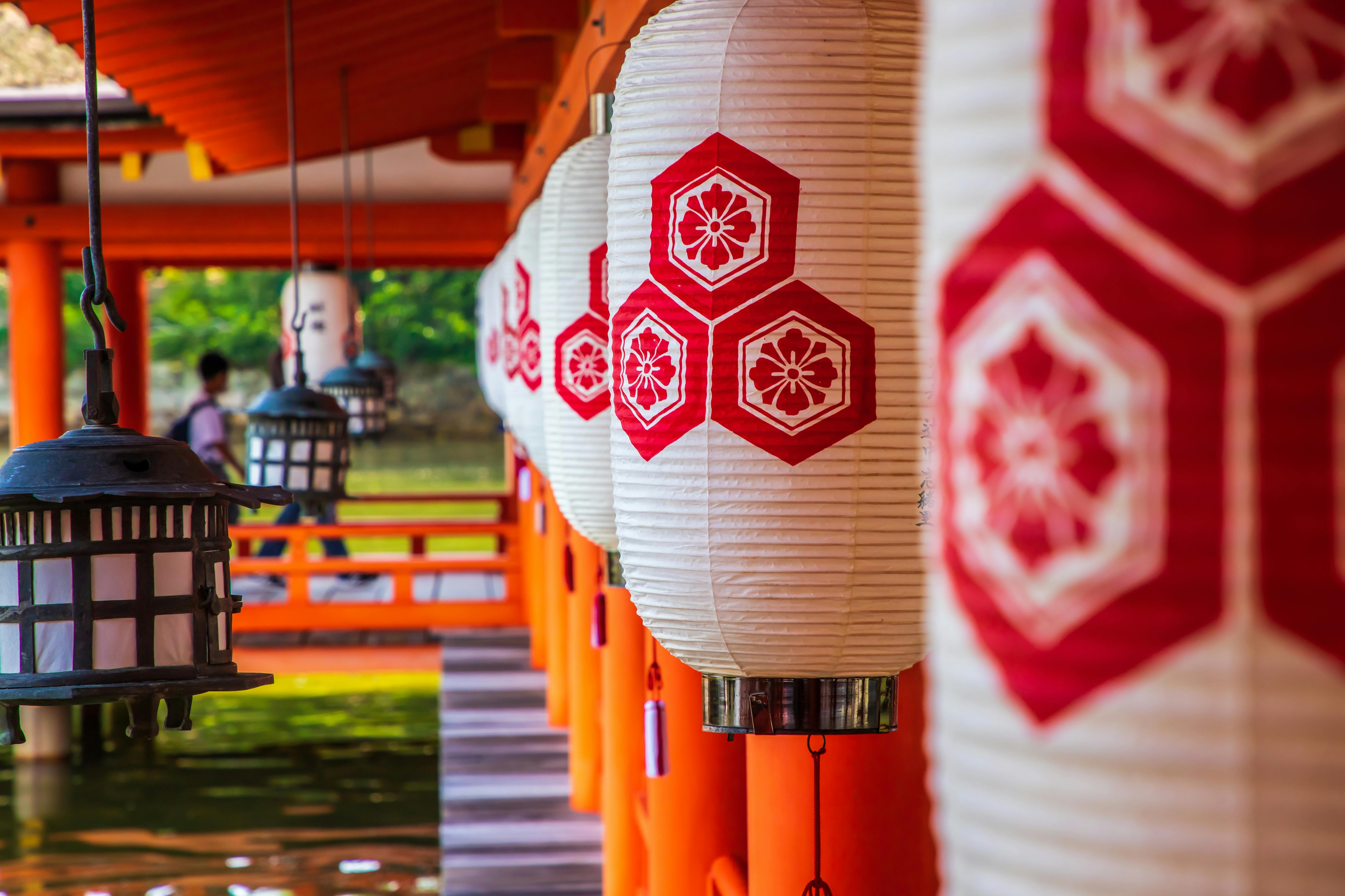 White Japanese lanterns with red hexagonal floral patterns hang in a row along a wooden corridor with orange pillars, near a body of water, with traditional lanterns and blurred figures in the background.