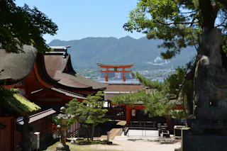 Traditional Japanese shrine buildings with curved roofs and a large red torii gate in the background, surrounded by trees and mountains under a clear blue sky.