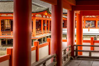 A view through red pillars of a traditional Japanese shrine with wooden walkways, railings, and elevated buildings featuring sloped roofs and detailed architectural elements.