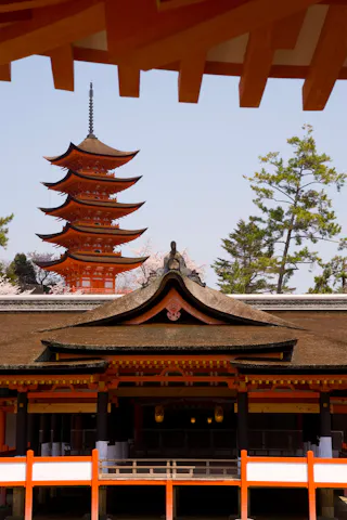 A vibrant red five-story pagoda stands behind a traditional Japanese temple with a curved roof, surrounded by green trees under a clear sky. Orange structural beams frame the top of the image.