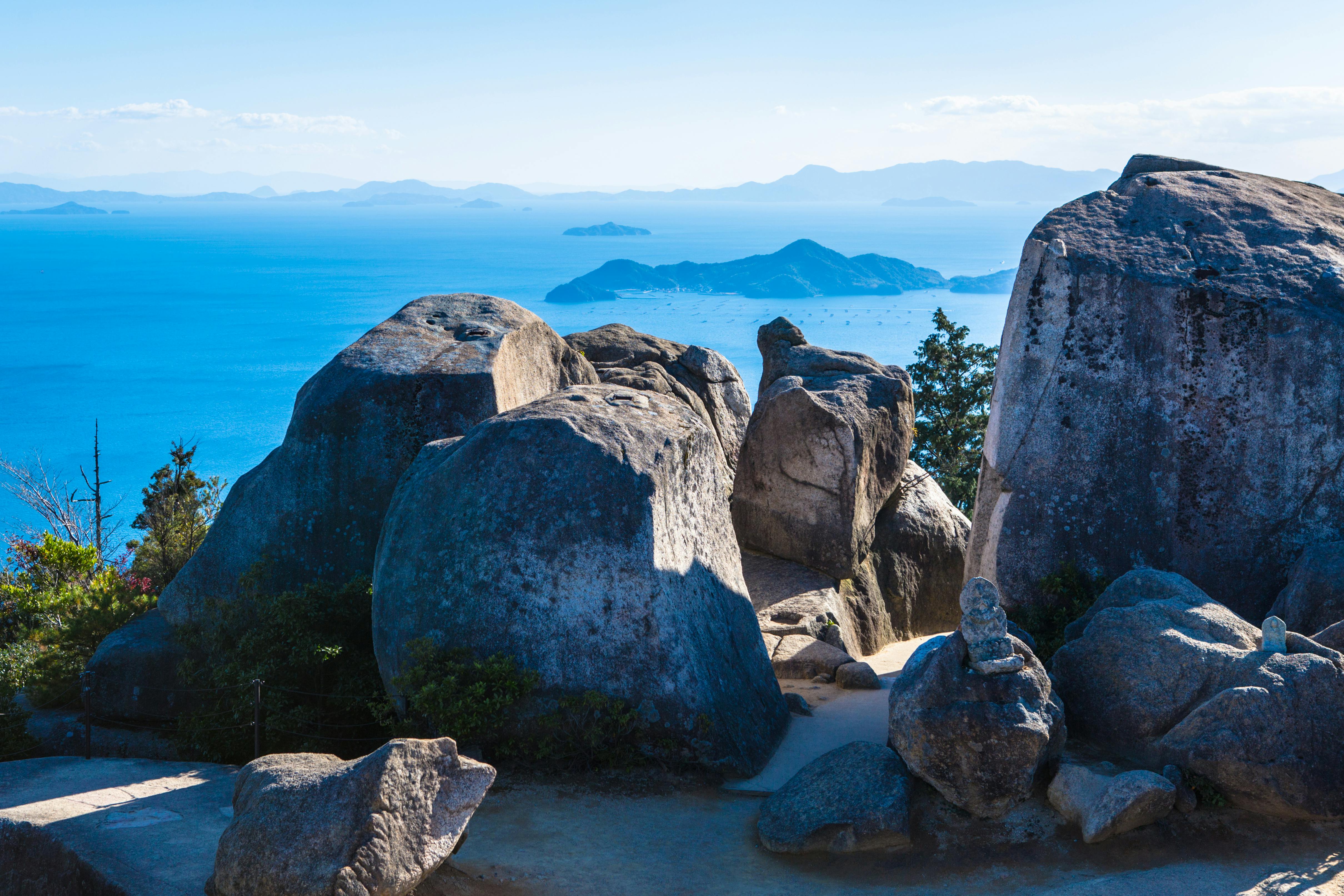 Large gray boulders sit atop a hill overlooking a blue sea, with distant islands and hazy mountains visible on the horizon under a clear sky. Some greenery and a sandy path surround the rocks.