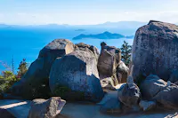 Large gray boulders sit atop a hill overlooking a blue sea, with distant islands and hazy mountains visible on the horizon under a clear sky. Some greenery and a sandy path surround the rocks.