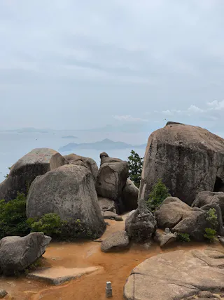 Large boulders and rocks rest on a dirt path with patches of green shrubs. In the background, distant mountains and islands rise above a hazy, overcast sky and calm sea.