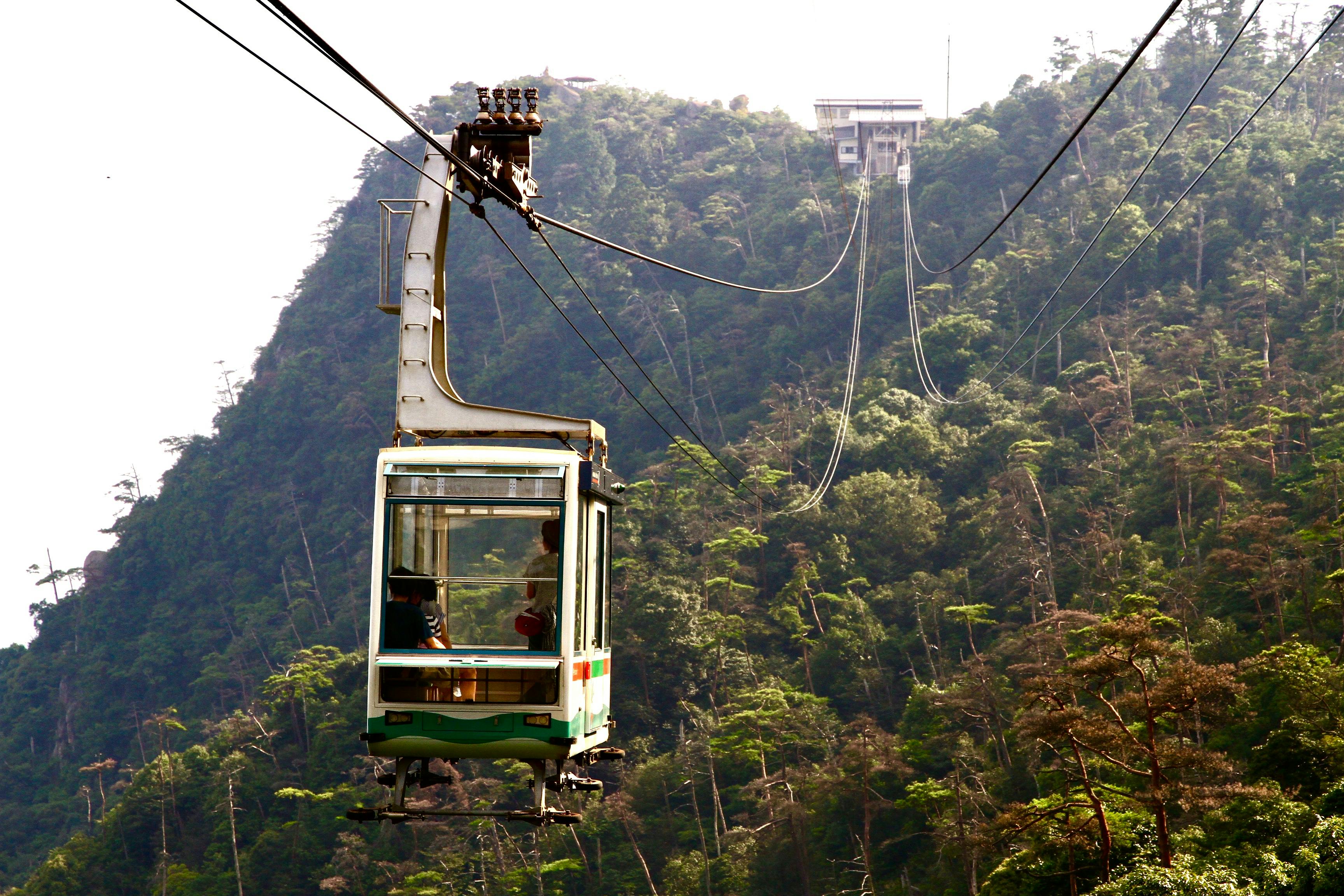 A cable car travels up a green, forested mountain toward a station at the summit, suspended on cables above the trees with people visible inside the cabin.