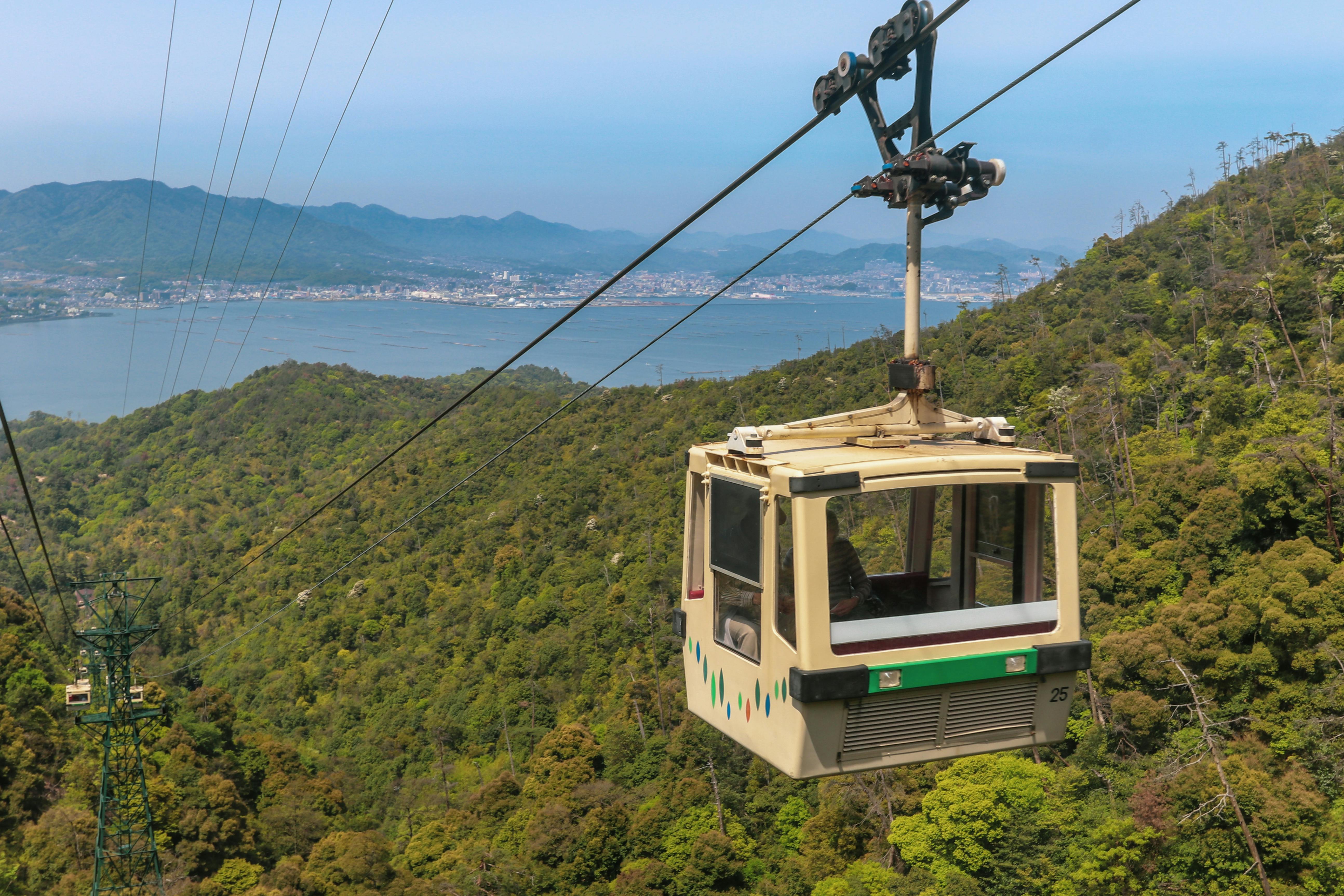 A cable car travels above a lush, green mountainside with a large body of water and cityscape visible in the background under a clear blue sky.