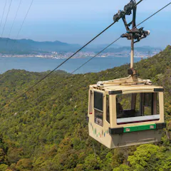 Miyajima Ropeway A cable car travels above a lush, green mountainside with a large body of water and cityscape visible in the background under a clear blue sky.