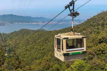 Miyajima Ropeway A cable car travels above a lush, green mountainside with a large body of water and cityscape visible in the background under a clear blue sky.