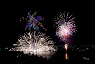 Colorful fireworks burst in the night sky above a body of water, reflecting vibrant reds, purples, and whites, with city lights faintly visible in the background.