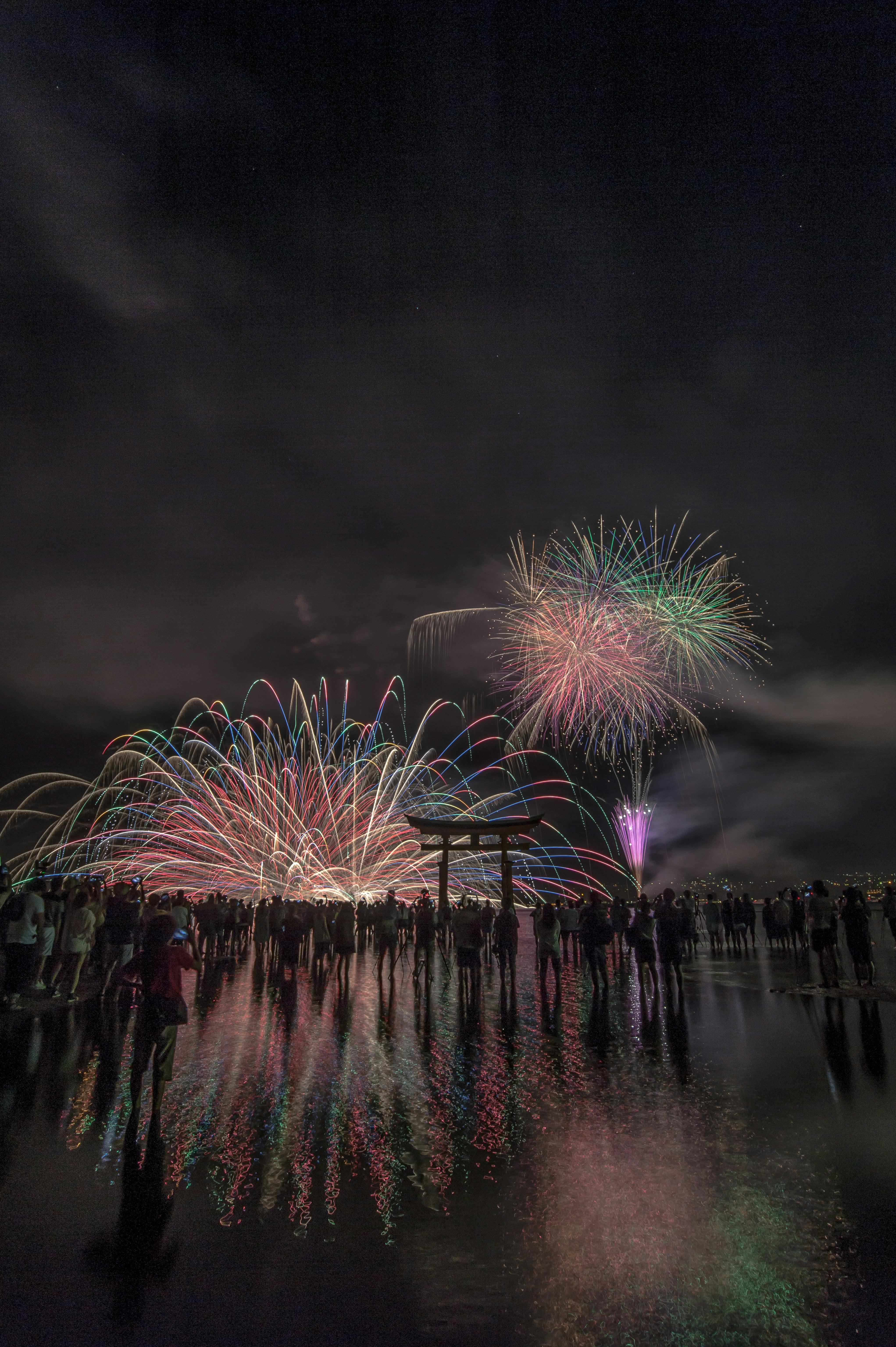 A crowd stands by the water at night, watching colorful fireworks burst and streak across the sky, their reflections shimmering on the wet surface below.