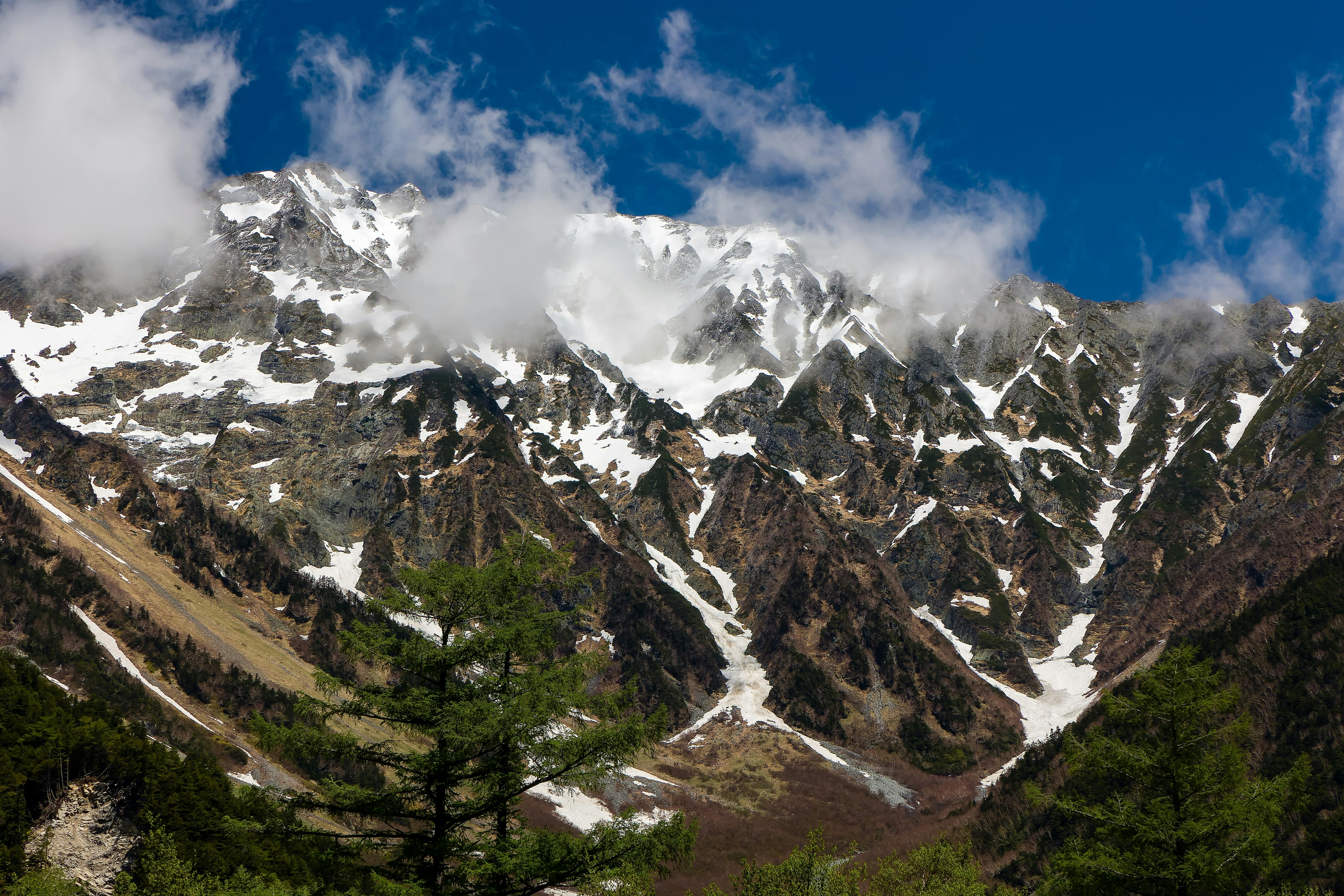 Snow-capped mountain peaks with patches of clouds under a blue sky, and green trees in the foreground.
