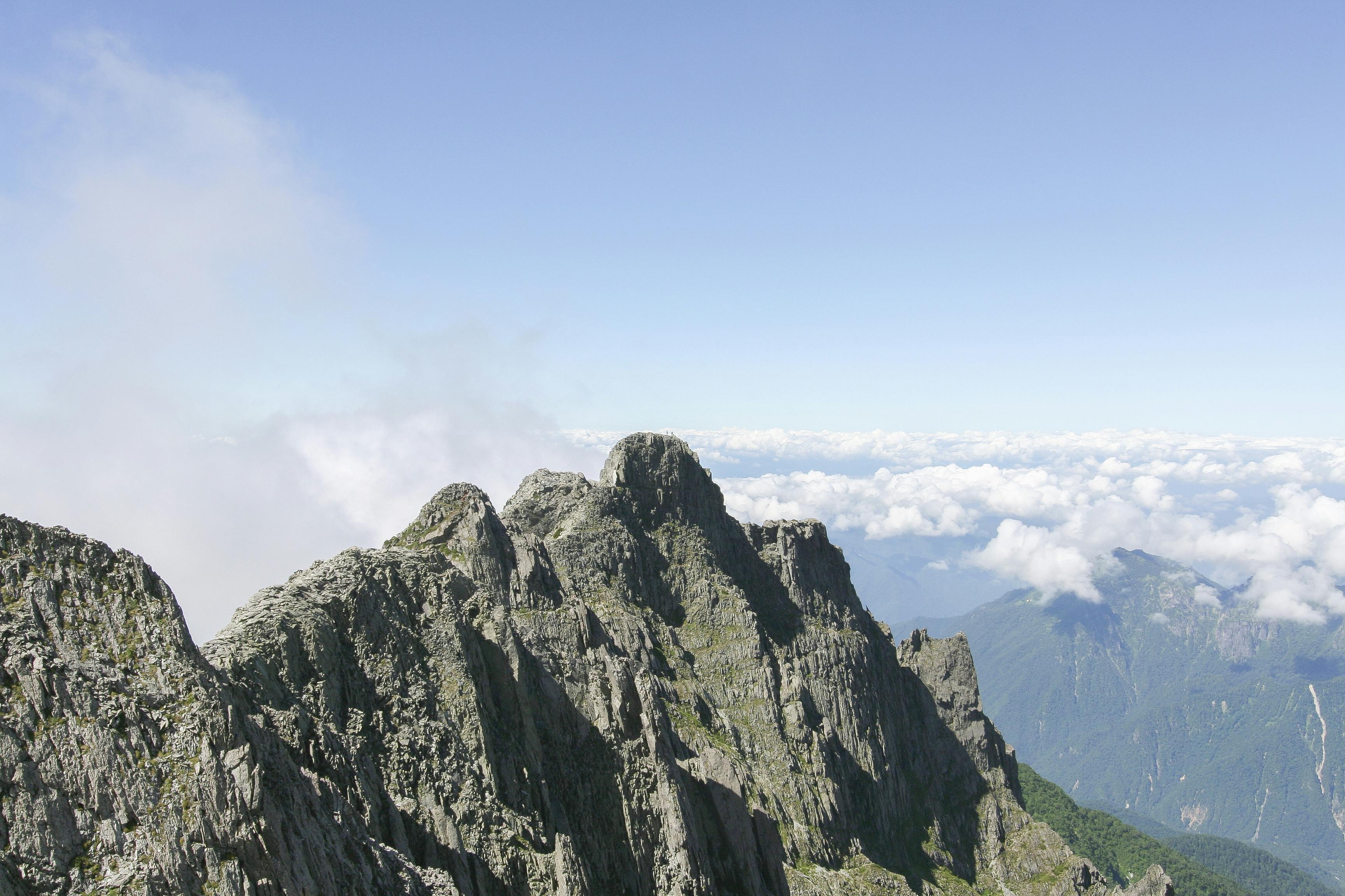 Jagged rocky mountain peaks stand against a clear blue sky with scattered clouds below, overlooking distant green hills and valleys.