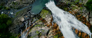Aerial view of a waterfall cascading over rocky cliffs surrounded by lush green vegetation, with clear water pools and streams visible below.