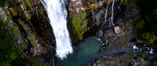 Aerial view of a tall waterfall cascading down rocky cliffs into a clear, emerald-green pool, surrounded by lush greenery and moss-covered rocks.