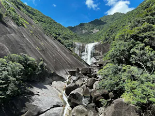 A waterfall cascades down smooth, steep rock cliffs surrounded by lush green trees and mountains under a bright blue sky with scattered clouds. Large boulders fill the foreground.