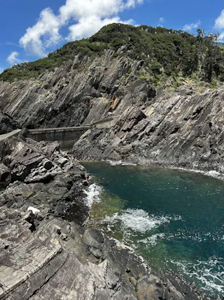 A rocky coastal pool with clear blue water surrounded by jagged, layered cliffs and green vegetation under a bright blue sky with scattered clouds.