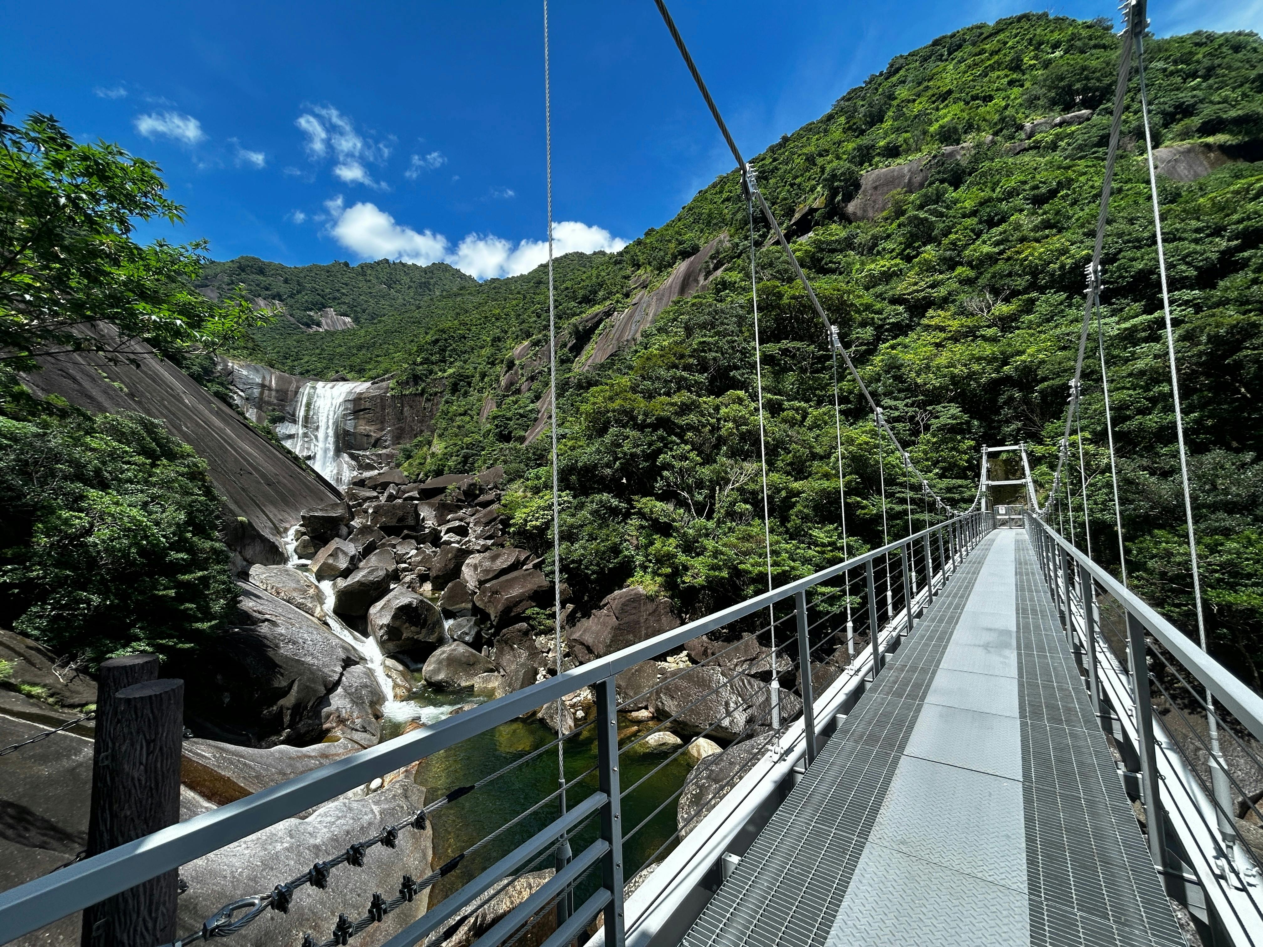A metal suspension bridge stretches toward a lush, green mountain with a tall waterfall cascading down rocky cliffs under a bright blue sky with scattered clouds.