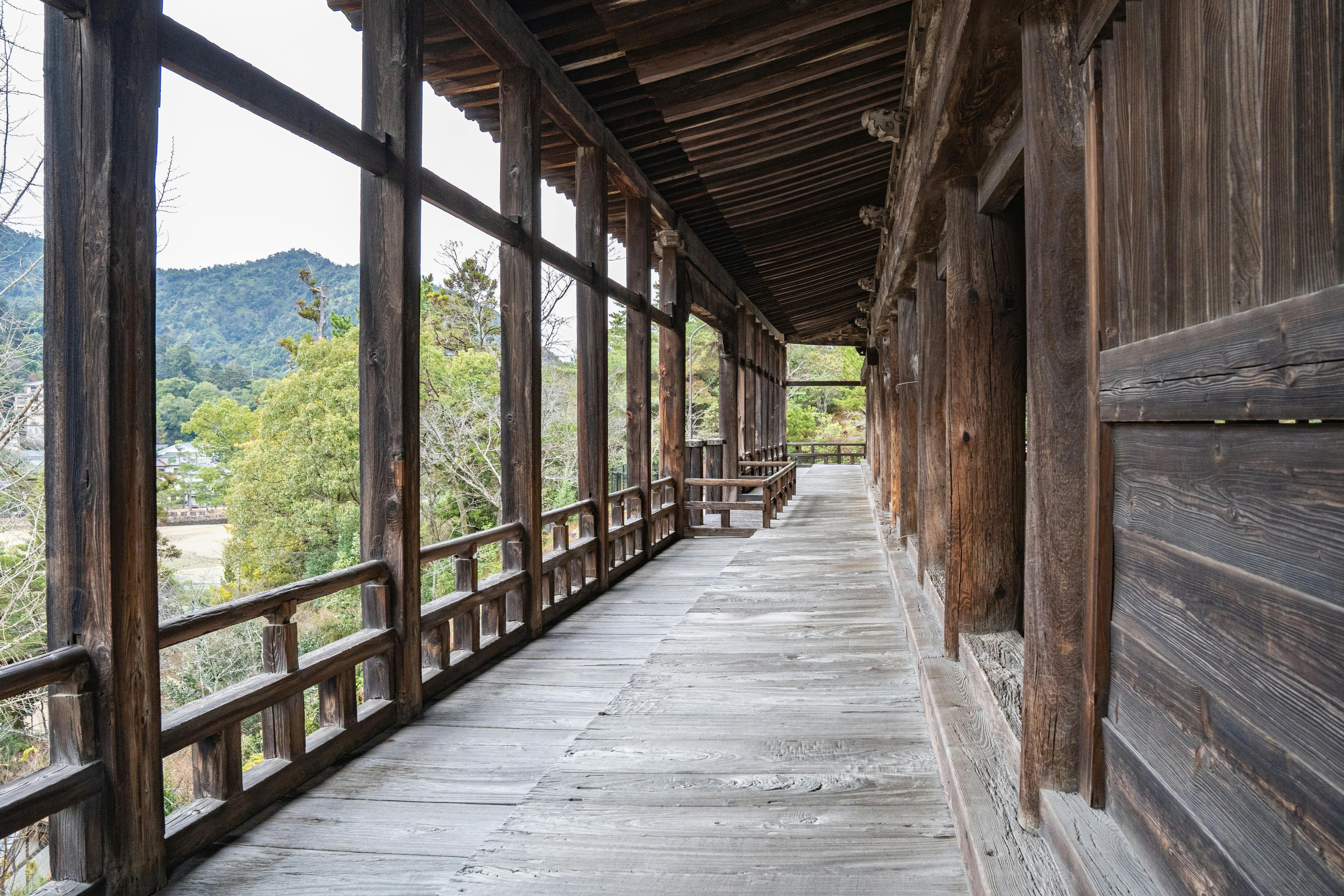 A wooden corridor with open sides reveals a scenic view of trees and distant mountains, with rustic railings and a weathered floor, typical of traditional Japanese architecture.
