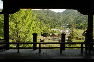 A person stands on a wooden balcony, taking a photo of a lush green landscape with trees, rooftops, and distant forested mountains under a partly cloudy sky.