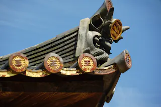 Close-up of an ornate roof corner on a traditional East Asian building, featuring decorative tiles with gold accents and intricate carvings, set against a clear blue sky.