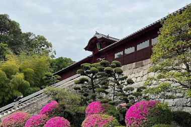 Traditional Japanese building with a sloped stone wall, surrounded by manicured trees and vibrant pink azalea bushes, under a cloudy sky.