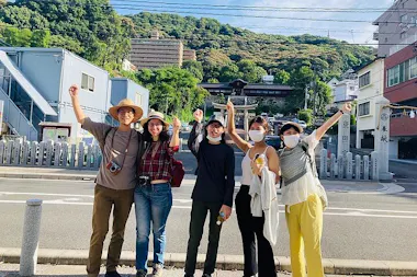Five people stand together on a street in front of a hilly, green area with buildings, smiling and raising their arms. Some wear hats and masks, suggesting a casual outing on a sunny day.