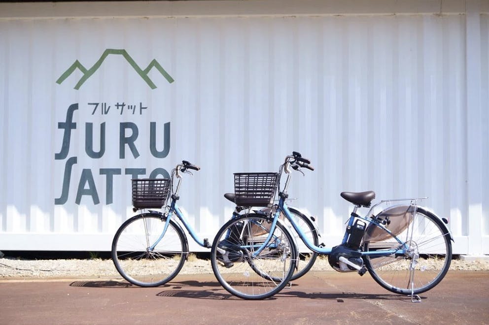 Two blue bicycles with front baskets are parked in front of a white wall with the "furu SATTO" logo and stylized green mountain graphic. Japanese text is also visible above the logo.