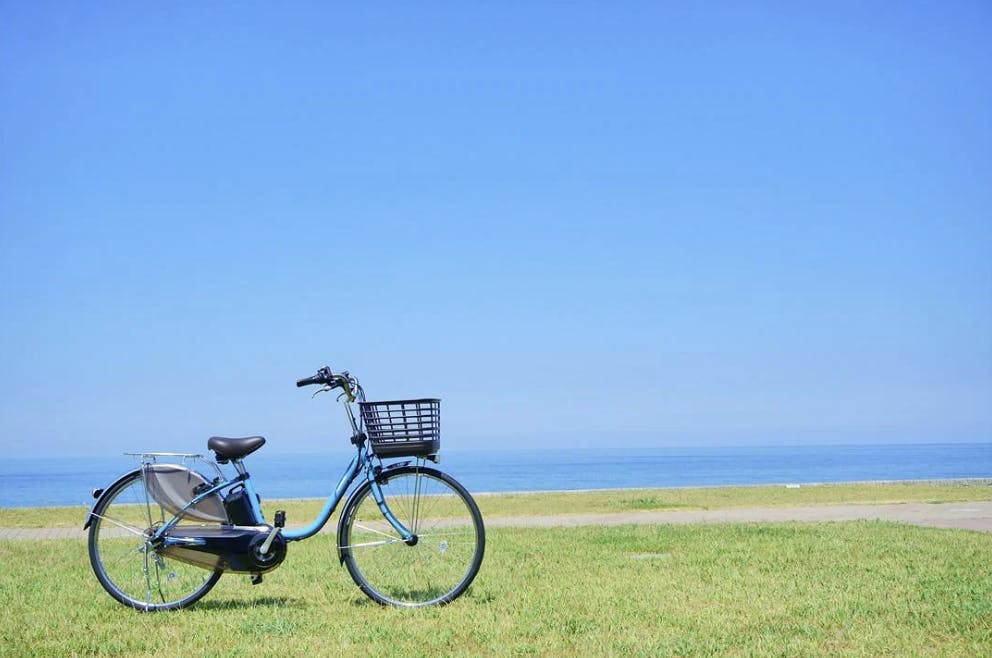 A blue bicycle with a black basket is parked on green grass near a paved path, with the ocean and a clear blue sky in the background.