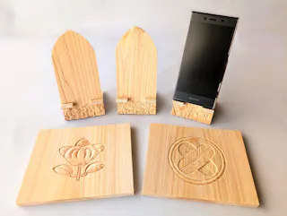 Two wooden phone stands with floral carvings hold a smartphone upright. In front, two square wooden plaques display intricate carvings: one of a flower, the other with a circular geometric design.