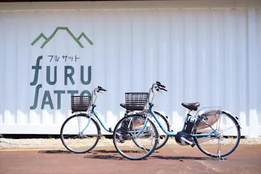 Two blue bicycles with baskets are parked in front of a white wall with green mountain graphics and the word "furusat" in English and Japanese writing.