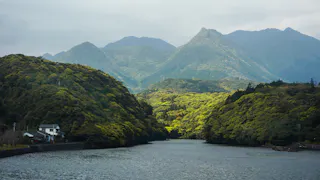 A river flows between lush green hills with a small white house on the left bank. Behind the hills, tall, forested mountains rise under a cloudy sky, creating a serene and scenic landscape.